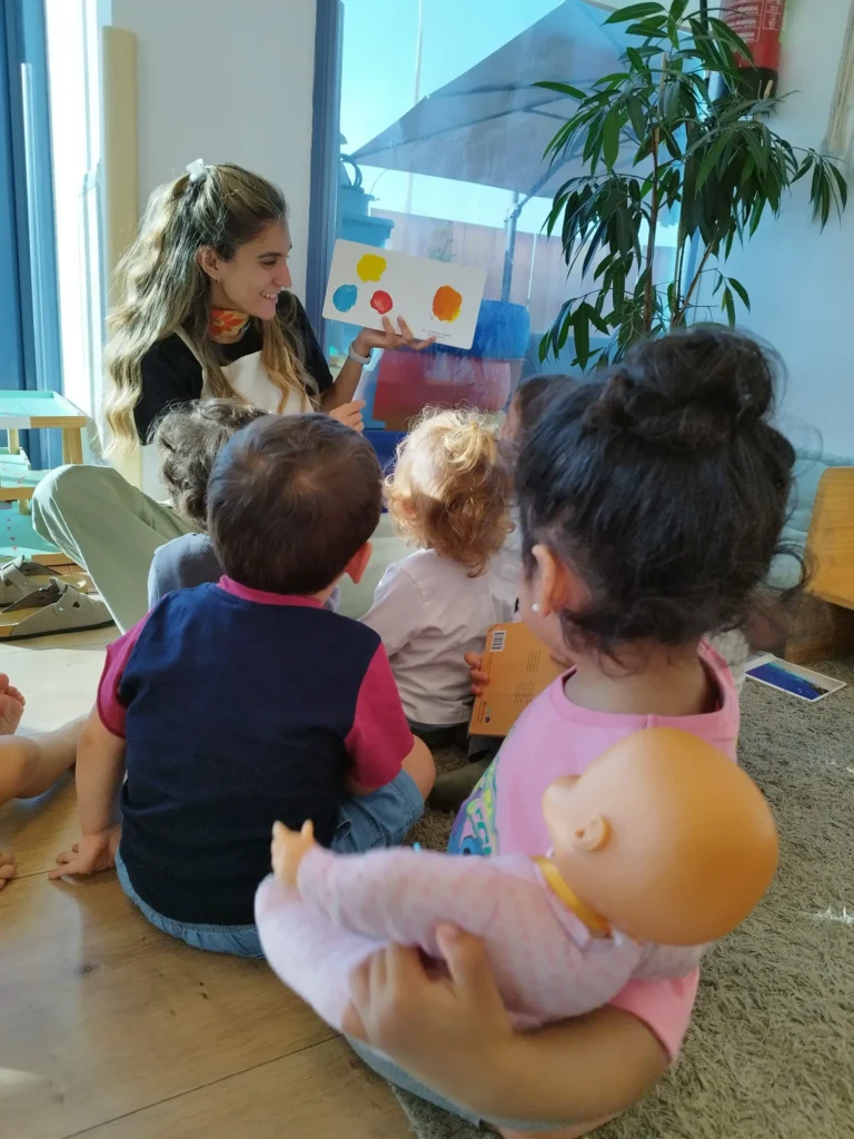 Grupo de niños de Comunidad Infantil sentados en el suelo prestando atención a una guía Montessori que les lee o presenta un material gráfico.